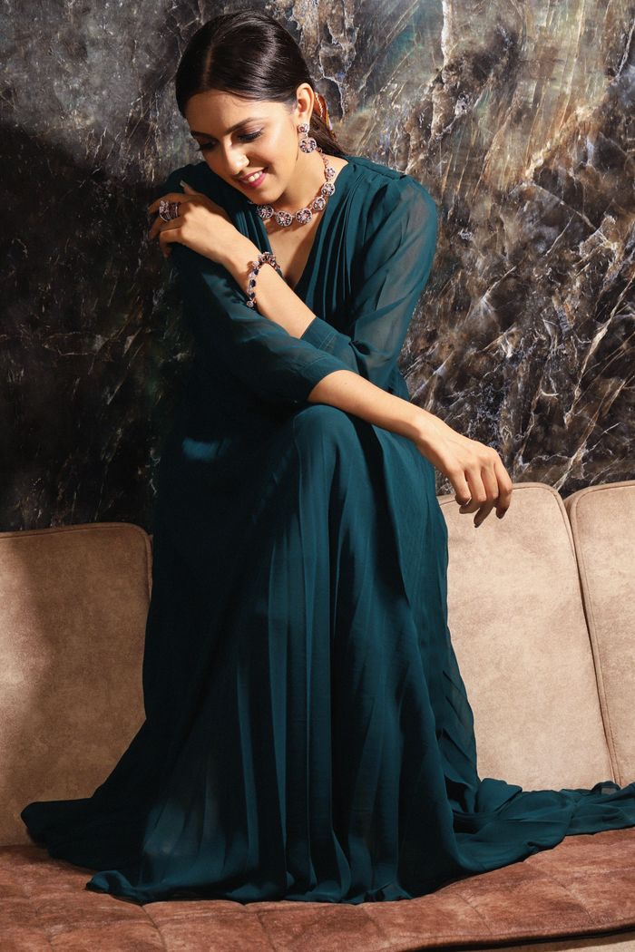 Elegant woman in a blue dress sitting and smiling indoors, showcasing modern style and jewelry.